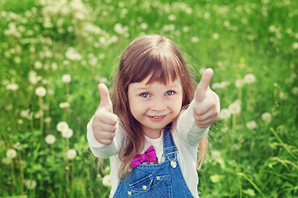 Little girl standing in green field giving two thumbs up to the camera.